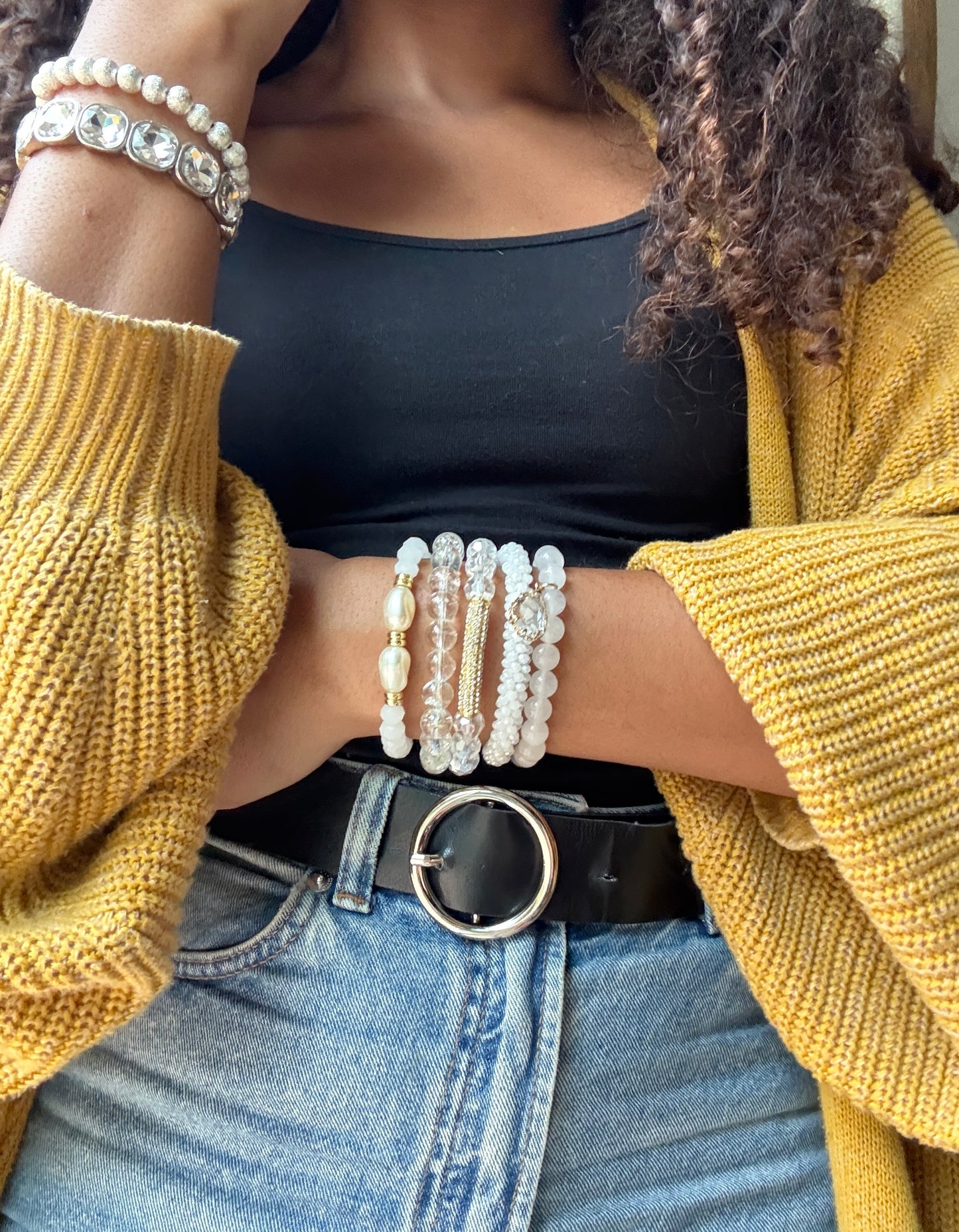 Person wearing a yellow sweater, black top, and blue jeans with multiple pearl bracelets on a blurred background.