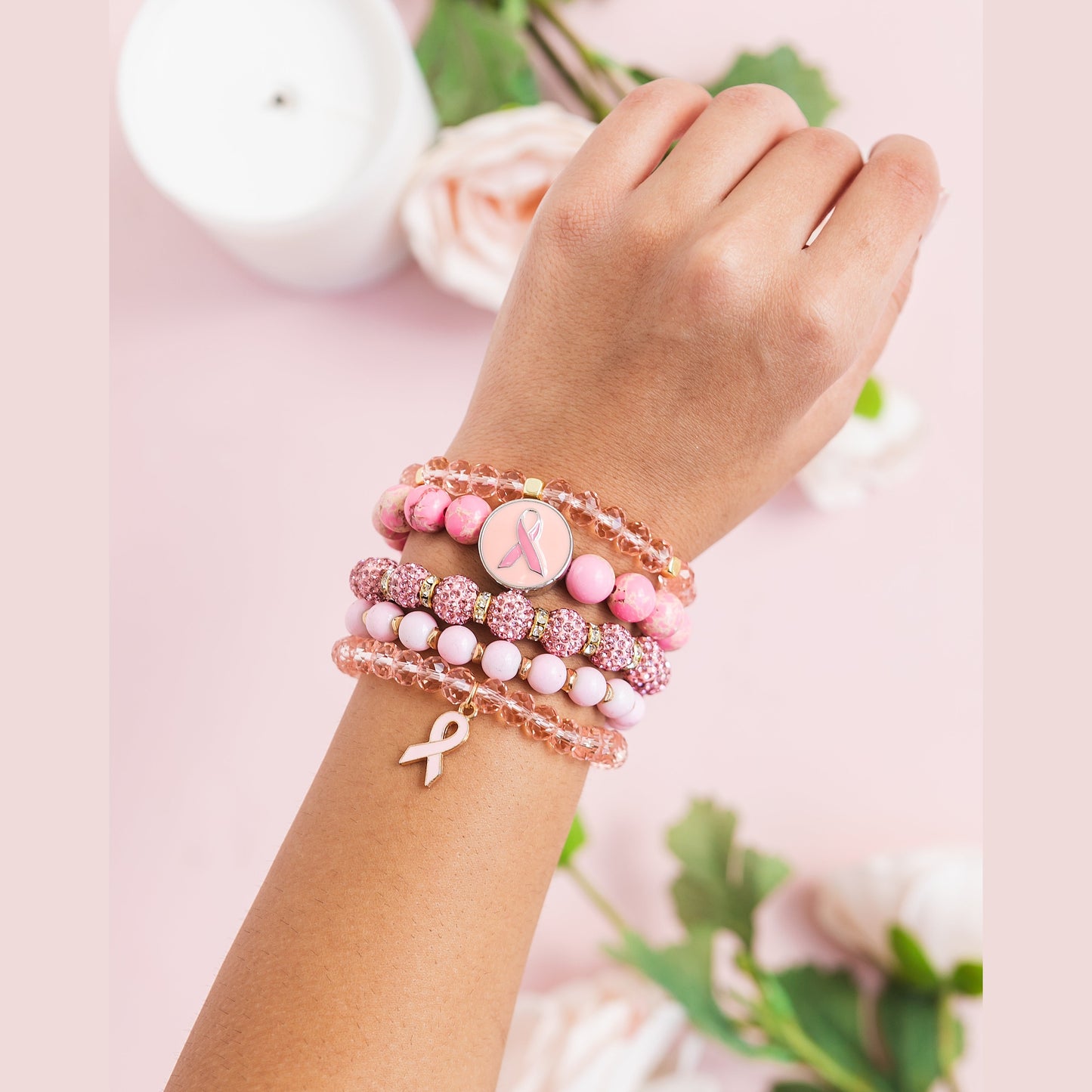 Hand wearing multiple pink beaded bracelets with breast cancer awareness ribbons on a soft pink background.