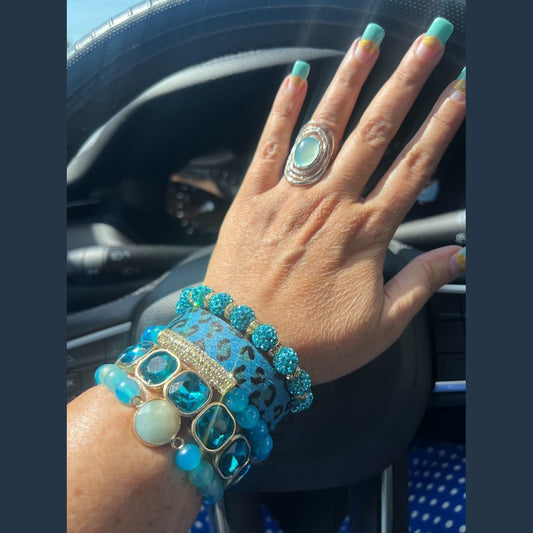 Hand with turquoise nail polish and jewelry, including a ring and bracelets, against a car interior background.