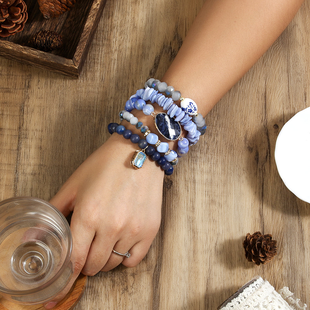 Hand wearing multiple blue beaded bracelets on a wooden surface with a glass of water and pine cones.