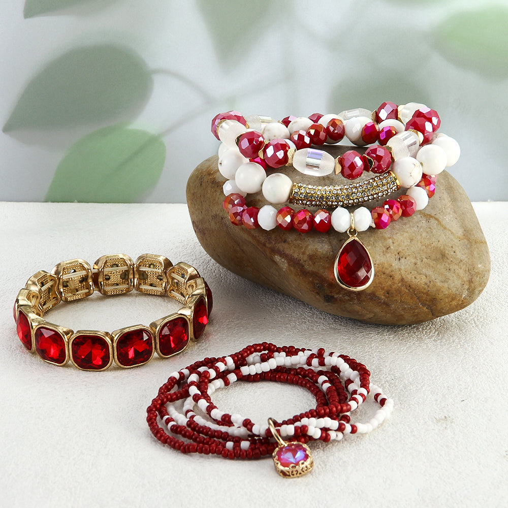 Jewelry set with red and white beaded bracelets on a stone surface with a blurred green leaf background.