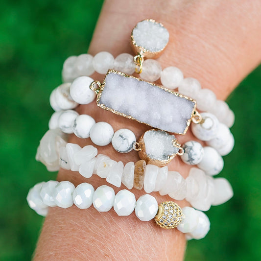 Stack of white beaded bracelets with gold accents on a wrist against a green background
